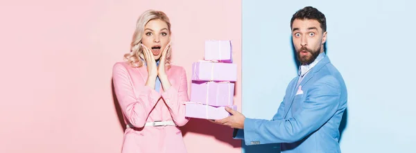 Tiro panorâmico de homem bonito dando presentes à mulher chocada no fundo rosa e azul — Fotografia de Stock