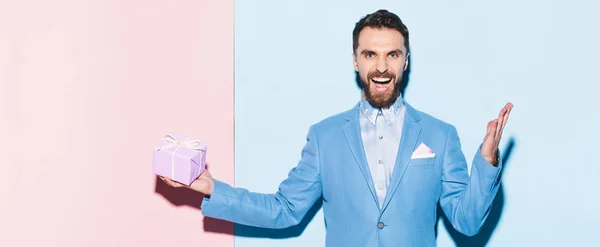 Panoramic shot of handsome and smiling man holding gift on blue and pink background — Stock Photo