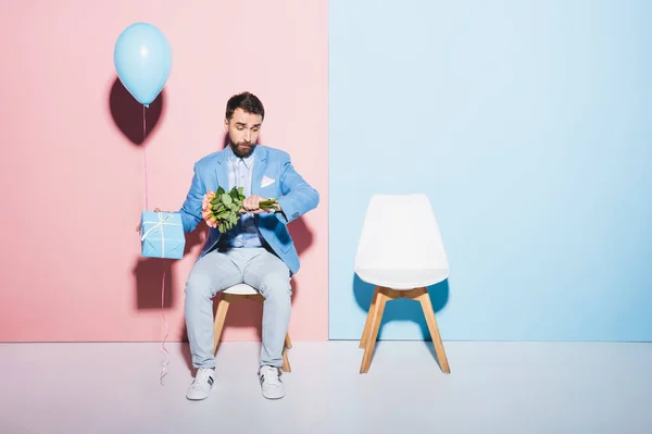 Uomo bello e nervoso guardando orologio da polso e tenendo palloncino, regalo e su bouquet sfondo blu e rosa — Foto stock