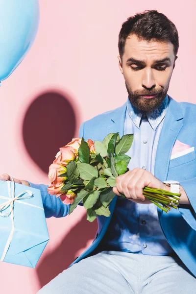 Handsome and nervous man looking at wristwatch and holding balloon, gift and on bouquet blue and pink background — Stock Photo