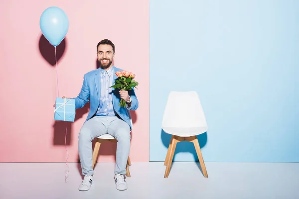 Hombre guapo y sonriente mirando reloj de pulsera y sosteniendo globo, regalo y en ramo de fondo azul y rosa - foto de stock