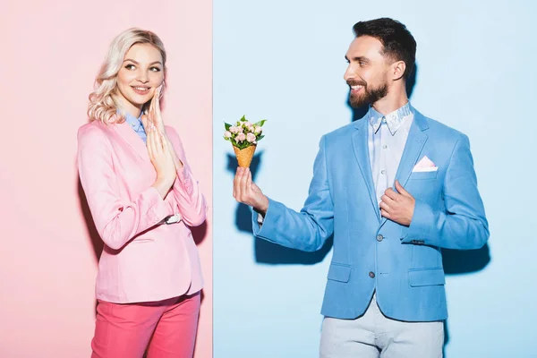 Homem sorrindo dando buquê à mulher sonhadora no fundo rosa e azul — Fotografia de Stock