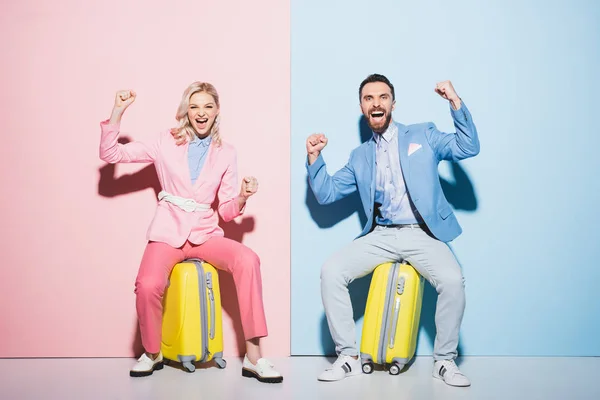 Mujer sonriente y hombre guapo sentado en bolsas de viaje y mostrando sí gesto sobre fondo rosa y azul - foto de stock