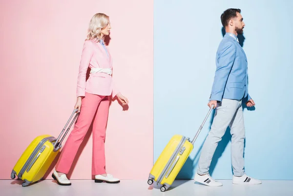 Seitenansicht einer attraktiven Frau und eines gutaussehenden Mannes, die mit Reisetaschen auf rosa und blauem Hintergrund gehen — Stockfoto