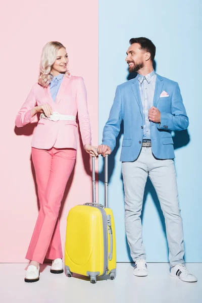 Улыбающаяся женщина и красивый мужчина с дорожной сумкой на розовом и синем фоне — стоковое фото