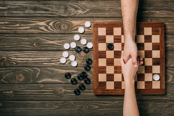 Cropped view of man and woman shaking hands above checkerboard with checkers on wooden background — Stock Photo