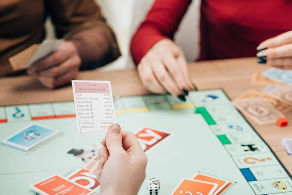 KYIV, UKRAINE - NOVEMBER 15, 2019: Cropped view of woman holding card by players and monopoly game on table — Stock Photo