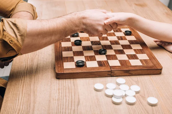 Cropped view of man and woman handshaking by checkerboard at table — Stock Photo