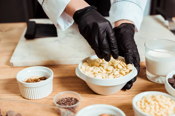 Vista ritagliato di cioccolatiere in guanti di lattice nero prendendo gocce di cioccolato bianco — Foto stock