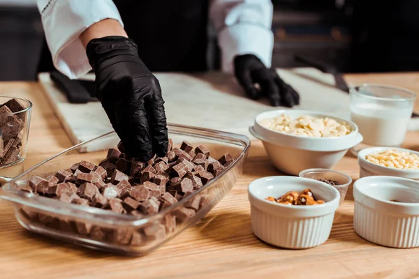 Обрізаний вид шоколатьє в чорних латексних рукавичках, що приймають темні шоколадні кубики — стокове фото