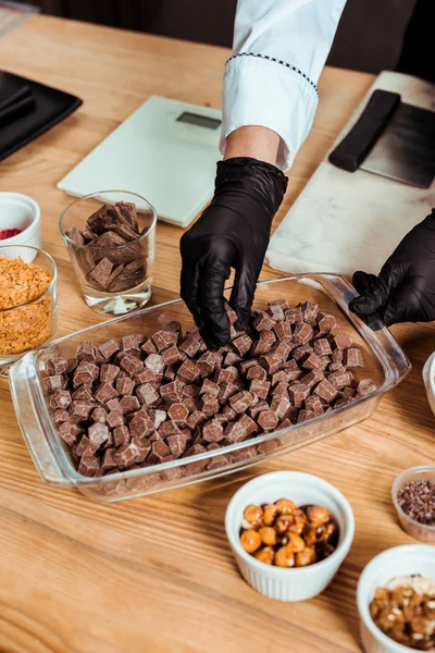 Ausgeschnittene Ansicht eines Chocolatiers in schwarzen Latex-Handschuhen, der dunkle Schokoladenwürfel nimmt — Stockfoto