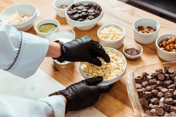 Ausgeschnittene Ansicht eines Chocolatiers in schwarzen Latex-Handschuhen, der weiße Schokoladenchips in der Nähe von Schüsseln nimmt — Stockfoto