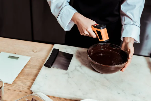 Vista recortada del chocolatero que sostiene el termómetro de cocina cerca del tazón con chocolate derretido - foto de stock