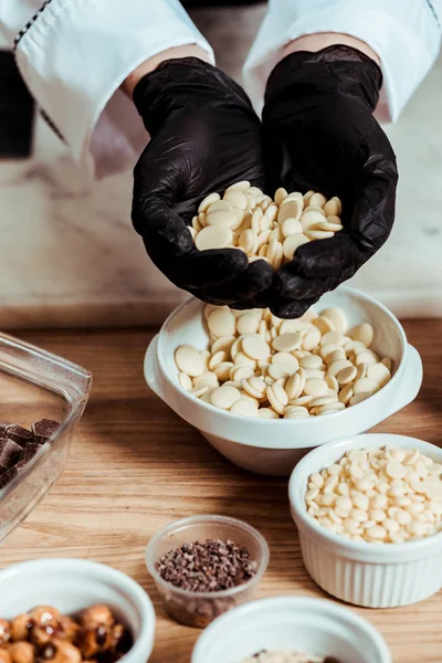 Vista recortada de chocolatero en guantes de látex negro con chips de chocolate blanco - foto de stock
