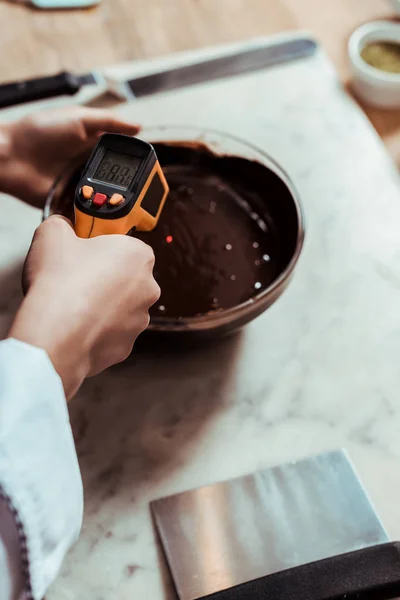 Abgeschnittene Ansicht des Chocolatiers, der das Kochthermometer in der Nähe der Schüssel mit geschmolzener dunkler Schokolade hält — Stockfoto