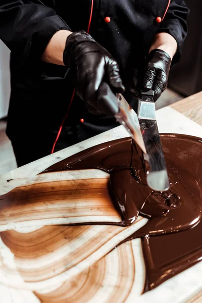 Cropped view of chocolatier holding cake scrapers near melted chocolate on surface — Stock Photo