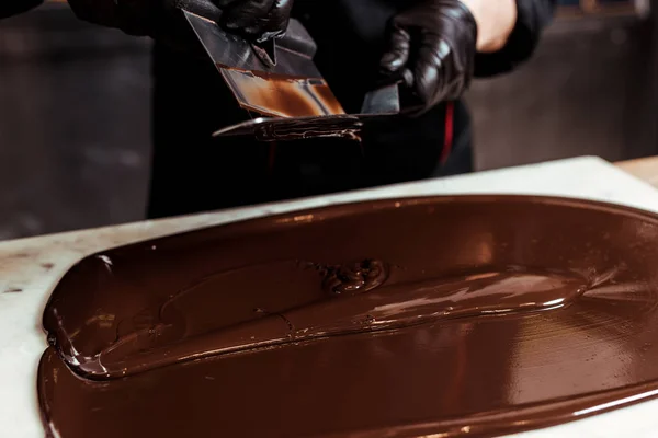 Cropped view of chocolatier holding cake scrapers near melted dark chocolate on surface — Stock Photo