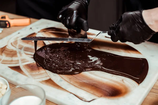 Vue recadrée de chocolatier tenant grattoir à gâteau près de chocolat noir fondu sur la surface — Photo de stock