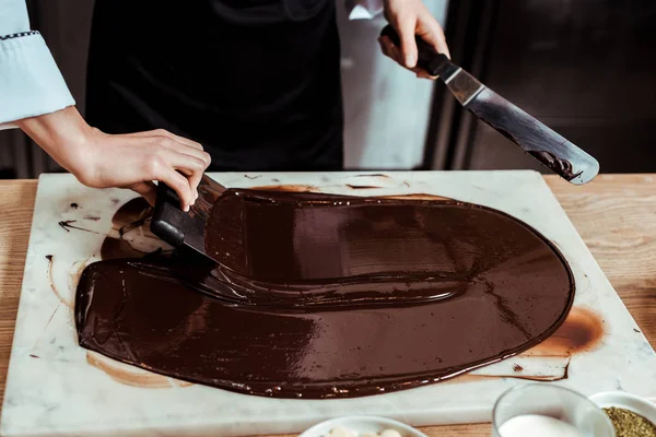 Vista cortada de chocolatier segurando raspadores de bolo perto de chocolate escuro derretido na superfície de mármore — Fotografia de Stock