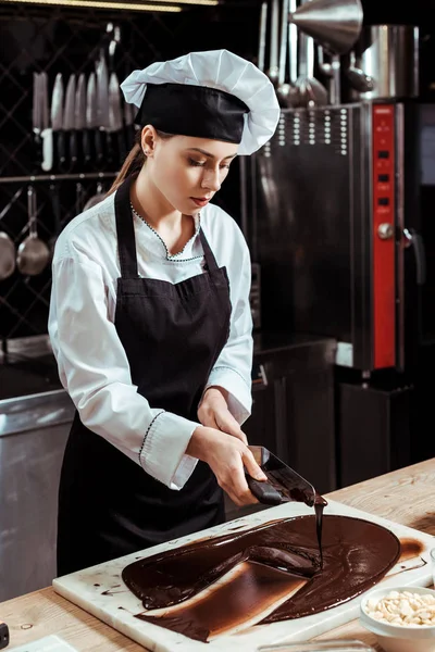 Attraktiver Chocolatier hält Kuchenschaber in der Nähe von geschmolzener dunkler Schokolade auf Marmoroberfläche — Stockfoto