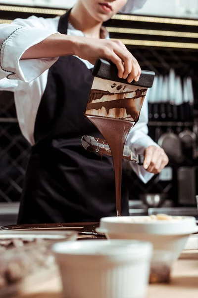 Cropped view of chocolatier holding cake scraper with melted dark chocolate pouring on surface — Stock Photo
