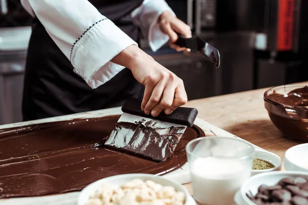 Abgeschnittene Ansicht des Chocolatiers, der Kuchenschaber in der Nähe von geschmolzener dunkler Schokolade und Milch hält — Stockfoto