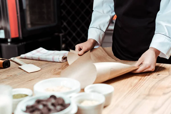 Cropped view of chocolatier holding baking paper near bowls with ingredients — Stock Photo