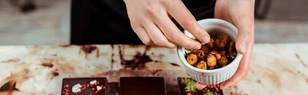 Panoramic shot of chocolatier holding bowl with caramelized hazelnuts near chocolate bars — Stock Photo