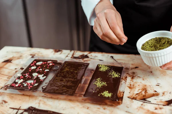 Vista recortada de chocolatero añadiendo polvo de pistacho en barra de chocolate negro - foto de stock