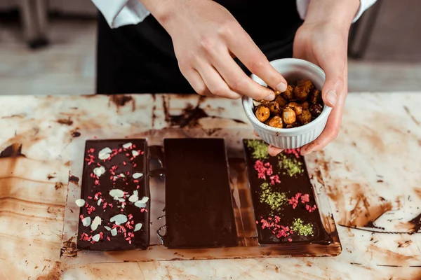 Cropped view of chocolatier holding bowl with caramelized hazelnuts near chocolate bars — Stock Photo