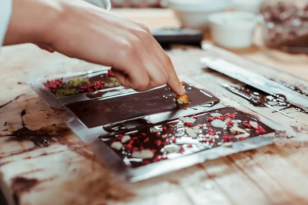 Vista recortada de chocolatero añadiendo avellanas caramelizadas en barra de chocolate negro - foto de stock