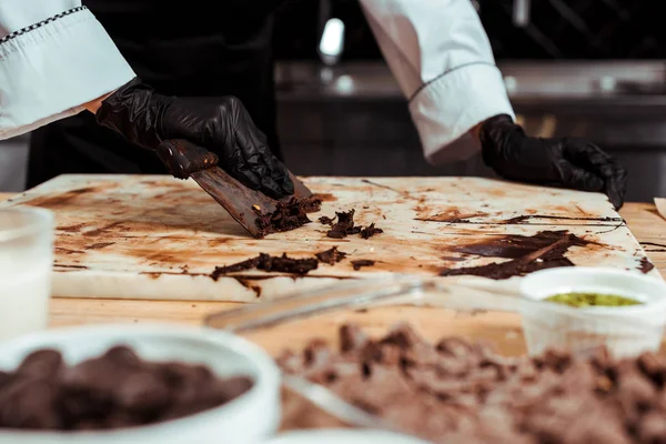 Ausgeschnittene Ansicht eines Chocolatiers in schwarzen Latex-Handschuhen, der dunkle Schokolade von der Oberfläche schabt — Stockfoto