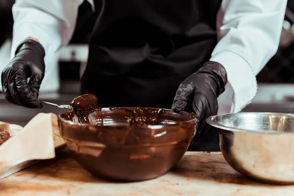 Vista recortada de chocolatero en guantes de látex negro sosteniendo batidor cerca de chocolate derretido en un tazón - foto de stock