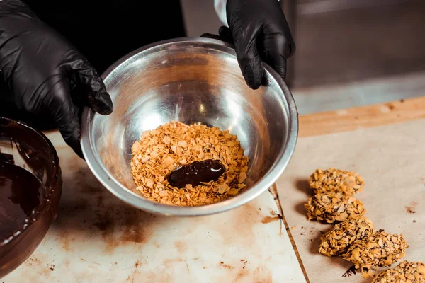 Vista cortada de chocolatier em luvas de látex preto segurando tigela com flocos perto de deliciosos doces de chocolate — Fotografia de Stock