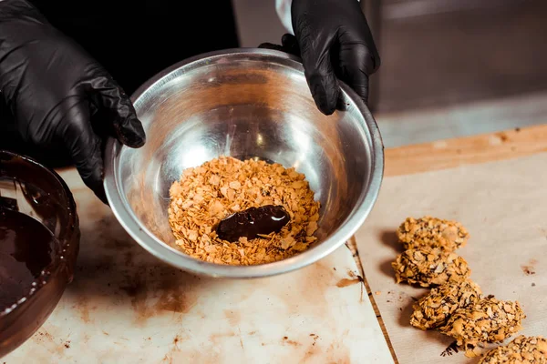 Vista ritagliata di cioccolatiere in guanti di lattice nero con ciotola con fiocchi vicino deliziosa caramella al cioccolato — Foto stock