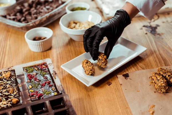 Ausgeschnittene Ansicht eines Chocolatiers in schwarzem Latex-Handschuh, der leckere Bonbons auf den Teller legt — Stockfoto