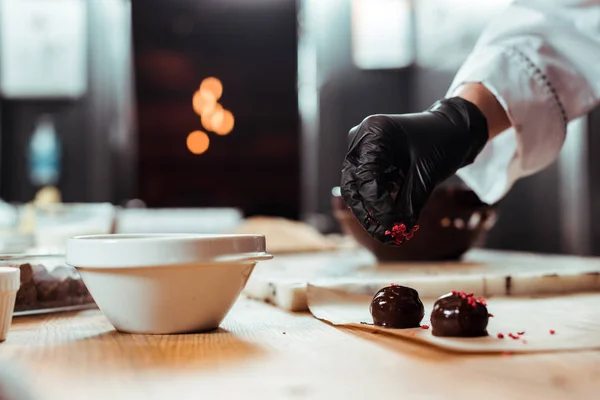 Vista recortada de chocolatero en guante de látex negro añadiendo frambuesas secas en caramelos frescos - foto de stock