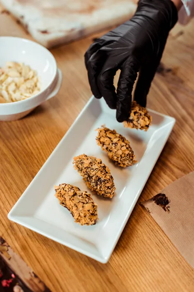 Cropped view of chocolatier in black latex glove taking delicious chocolate candy with flakes — Stock Photo