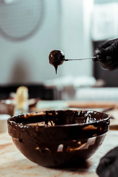 Vue recadrée du chocolatier en gant de latex noir tenant bâton avec de savoureux bonbons près du chocolat dans un bol — Photo de stock