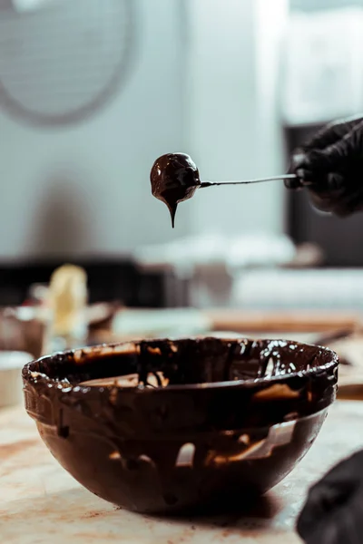 Cropped view of chocolatier in black latex glove holding stick with tasty candy near chocolate in bowl — Stock Photo