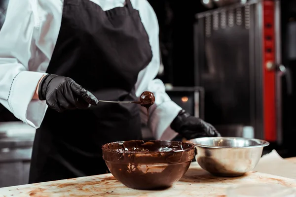 Vista recortada de chocolatero en delantal negro sosteniendo palo con dulces sabrosos cerca del chocolate en un tazón — Stock Photo