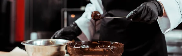 Plano panorámico de chocolatero en delantal negro sosteniendo palo con sabrosos dulces cerca de chocolate en tazón - foto de stock