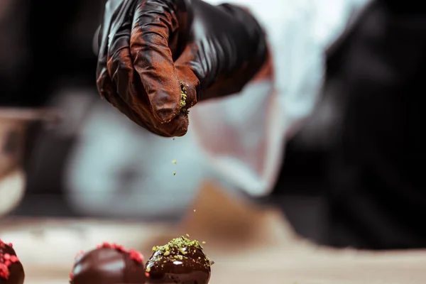 Обрезанный вид шоколада в черной латексной перчатке, добавляющий зеленый фисташковый порошок на свежие конфеты — стоковое фото