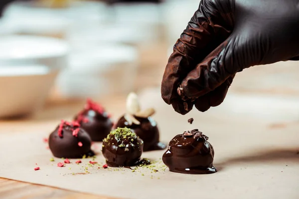 Vue recadrée du chocolatier en gant de latex noir ajoutant des copeaux de chocolat sur des bonbons frais — Photo de stock