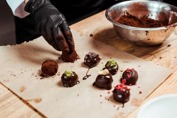 Cropped view of chocolatier in black latex glove preparing truffle candies near chocolate balls — Stock Photo
