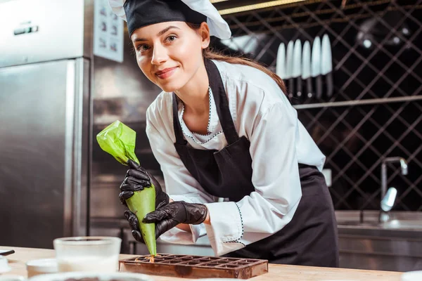 Messa a fuoco selettiva di cioccolatiere felice in guanti di lattice nero tenendo sacchetto di pasticceria vicino a stampi di cioccolato — Foto stock