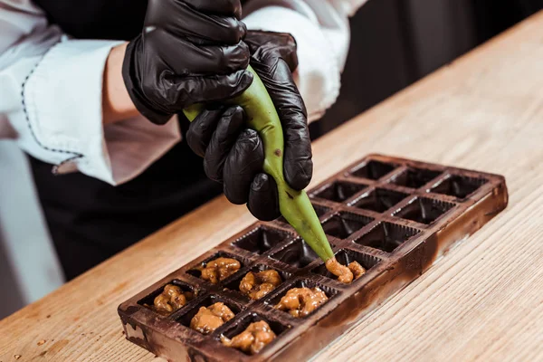Vista cortada de saco de pastelaria segurando chocolate com nozes caramelizadas perto de moldes de chocolate — Fotografia de Stock