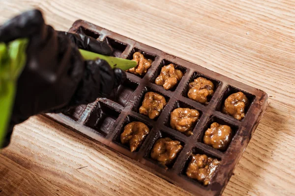 Vista cortada de chocolate em luvas de látex segurando saco de pastelaria com nozes caramelizadas perto de moldes de chocolate — Fotografia de Stock