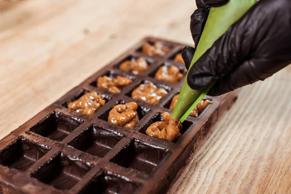 Vista cortada de chocolate em luva de látex segurando saco de pastelaria com nozes caramelizadas perto de moldes de chocolate — Fotografia de Stock