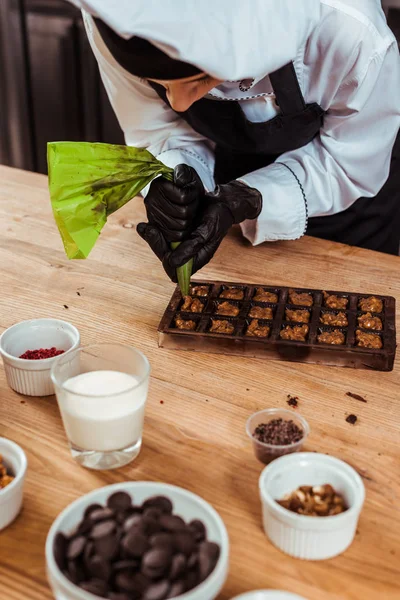 Chocolatier in Latexhandschuhen mit Teigtasche mit karamellisierten Nüssen in der Nähe von Schokoladenformen — Stockfoto